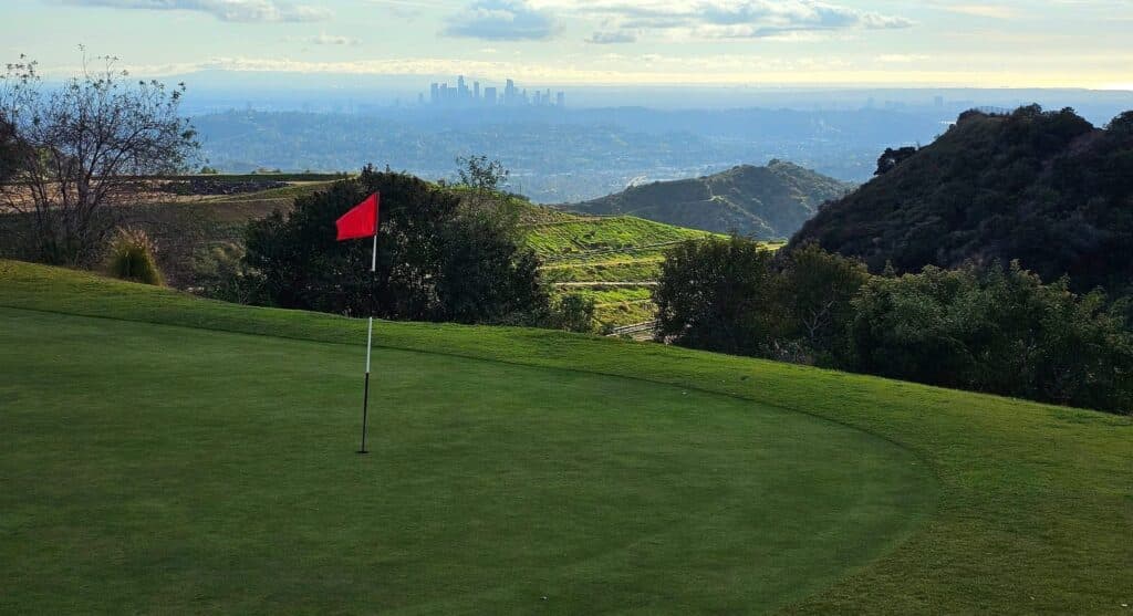 A red flag on a Scholl Canyon putting green with downtown Los Angeles visible beyond the San Rafael Hills. Photo courtesy Scholl Canyon Golf & Tennis Club (schollcanyongc.com). Used for editorial coverage.
